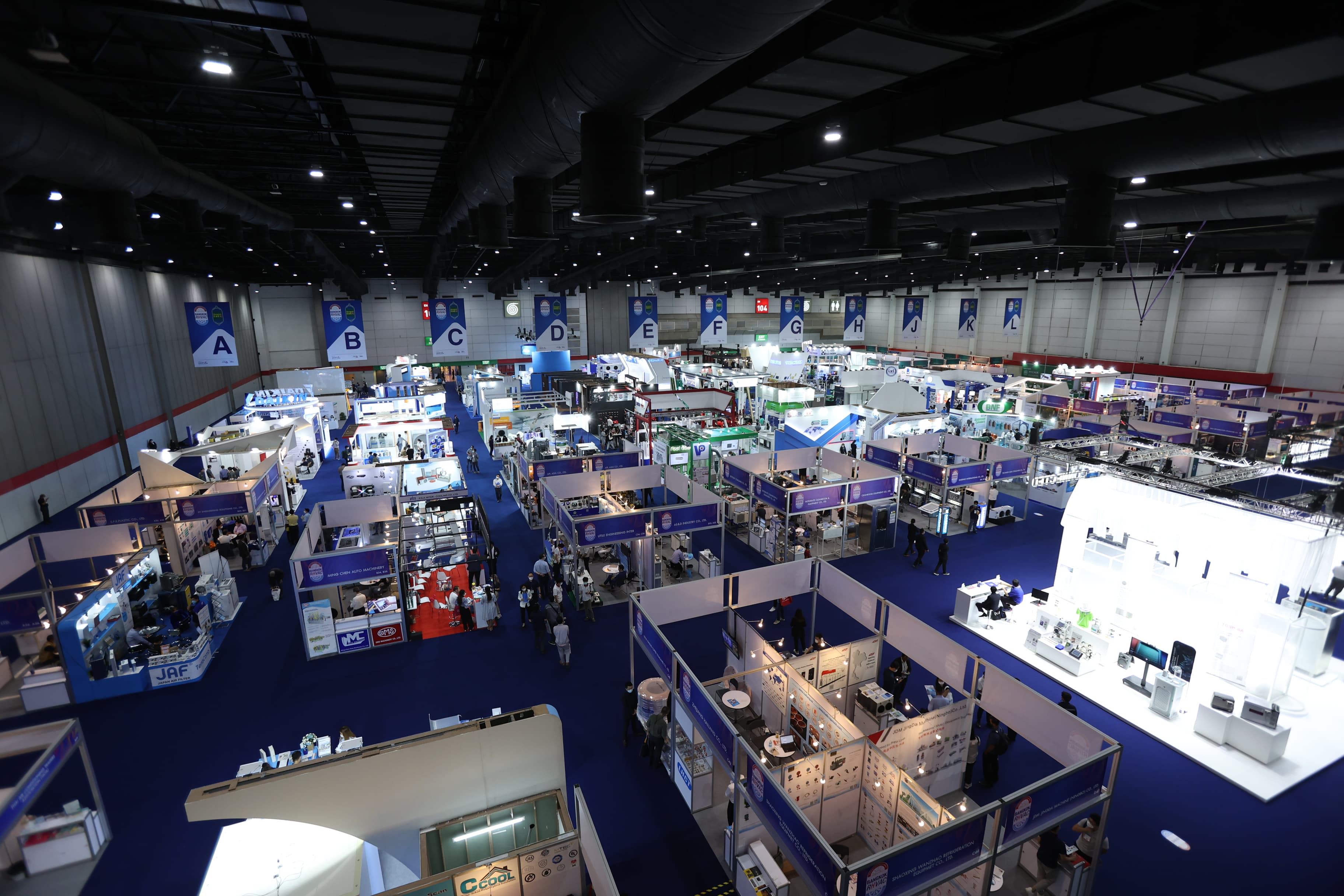 Large trade show floor with many exhibition booths, blue carpet, and hanging alphabetical banners.